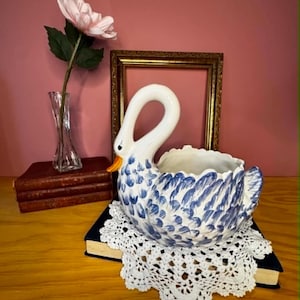 Peut inclure: Un pot de fleurs en forme de cygne en céramique blanche et bleue, avec un bec jaune, posé sur un napperon en dentelle blanche. Une fleur rose dans un vase en verre est en arrière-plan.