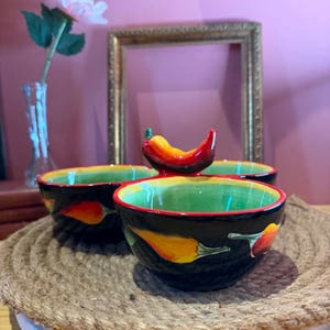 Peut inclure: Un bol de service en céramique à trois compartiments avec un extérieur noir orné de motifs de piments jaunes et rouges. L'intérieur est vert vif avec un bord rouge. Une poignée sculptée en forme de piment rouge et jaune est au centre.
