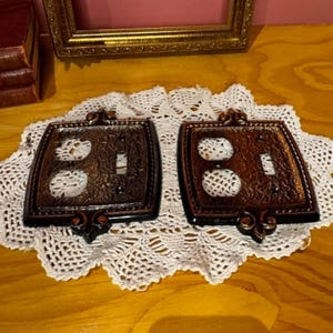 May include: Two decorative, ornate light switch covers. The covers are square with a dark brown finish and bronze accents. Each cover has a different configuration of holes for switches and outlets. They are displayed on a white lace doily.