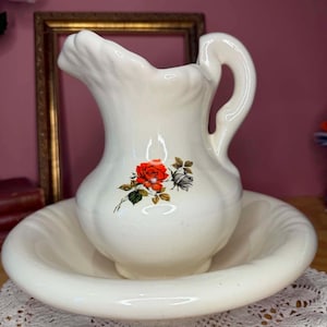 May include: A cream-colored ceramic pitcher and matching basin set. The pitcher features a scalloped rim, a curved handle, and a central floral decal depicting a vibrant red rose with green leaves and a smaller, muted grey rose bud.