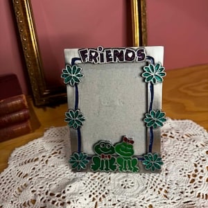 May include: A silver-colored picture frame with the word "FRIENDS" in purple letters. The frame is decorated with green flower accents and two cartoon frogs. The frame is sitting on a white lace doily.