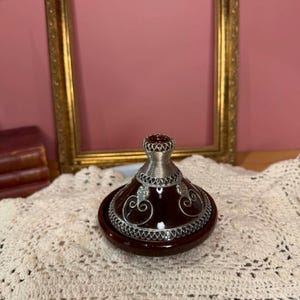 Peut inclure: Un couvercle de tajine en céramique marron foncé orné de superpositions en métal argenté. Le couvercle conique présente un bouton décoratif en métal au sommet et des motifs métalliques en volutes autour de la base et de la partie centrale.