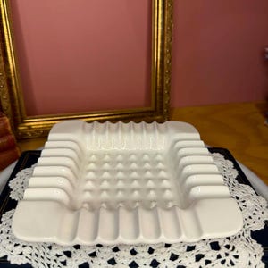 Peut inclure: Un cendrier carré en céramique blanche présentant une grille de bosses surélevées sur la base intérieure et une série de rainures profondes et arrondies sur les quatre bords, conçu pour maintenir des objets avec une finition lisse et brillante.