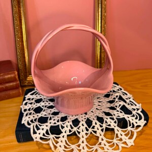 May include: A glossy, light pink ceramic basket featuring a smooth, curved bowl with a scalloped rim and a single, arched handle that extends from the sides. The surface of the ceramic is solid in colour with a reflective, polished finish throughout.