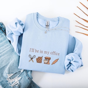 May include: A light blue crewneck sweatshirt featuring brown embroidery that reads "I'll be in my office" above icons of a whisk, an oven mitt, a stand mixer, and a rolling pin. The garment has long sleeves and a ribbed collar, neckline, and cuffs.