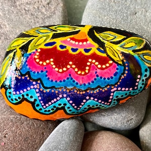 May include: A hand-painted rock with a colorful floral design. The rock is painted in shades of orange, yellow, red, pink, blue, and black. The design features a large flower with intricate details and leaves.
