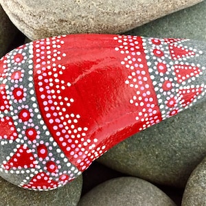 May include: A smooth, gray stone painted with red and white dots in a geometric pattern. The stone is painted red in the center and has a white and pink dotted border.