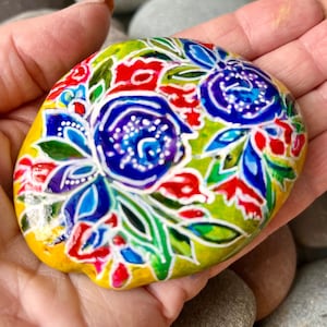 May include: Hand-painted rock with a floral design. The rock is yellow with blue, red, green, and white flowers.
