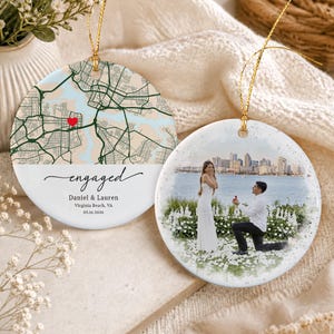 May include: Two round ceramic ornaments with gold hanging strings. One features a map with a red heart, the text "engaged", "Daniel & Lauren", "Virginia Beach, VA", and "05.16.2026". The other shows a photograph of a marriage proposal by a waterfront.
