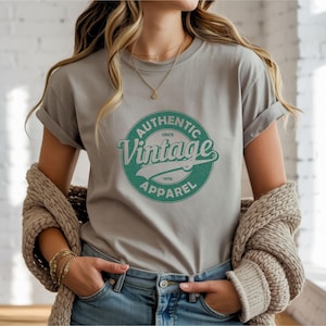 以下が含まれることがあります： 袖をまくったグレーのTシャツ。緑と白の円形グラフィックがあり、「Authentic Vintage Apparel Since 1976」と書かれています。Tシャツは柔らかい素材で作られています。