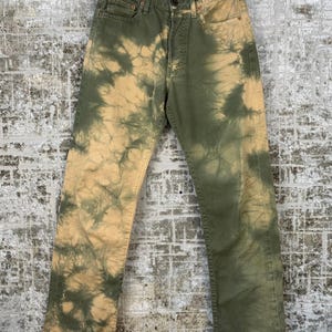 Op de afbeelding: Groene en bruine tie-dye jeans hangen aan een zwarte hanger. De jeans hebben een knoop- en ritssluiting, met zakken aan de voor- en achterkant. Het tie-dye patroon is een mix van groen en bruin.