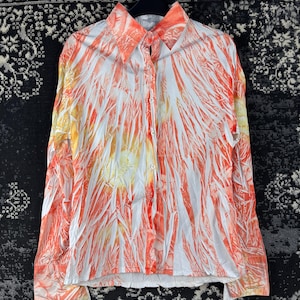 Può includere: Camicia abbottonata a maniche lunghe con un motivo astratto arancione e bianco. La camicia ha un colletto e polsini ed è appesa a una gruccia nera. Lo sfondo è un tessuto fantasia.