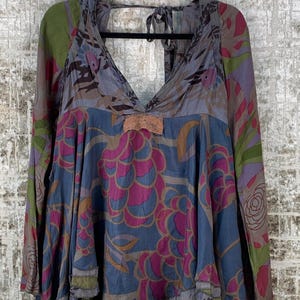 May include: A long-sleeved top with a deep V-neck and a flowing silhouette. The top features a mix of floral and abstract patterns in shades of blue, pink, green, and purple. A small lace detail accents the neckline. The garment is hanging on a black hanger.