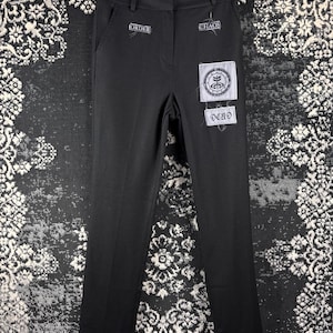 Può includere: Pantaloni neri con toppe che recitano "ORDER", "CHAOS" e "DEAD". Gli occhielli argentati sono presenti alle caviglie. Le toppe hanno uno sfondo bianco con testo e disegni neri. I pantaloni sono appesi a una gruccia nera.