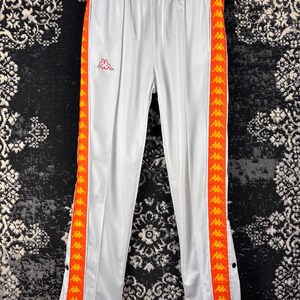 May include: White track pants with orange side stripes featuring a repeating logo pattern. The pants have a drawstring waist and snap closures at the ankles. A small logo is visible on the left leg.