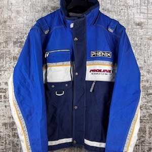 May include: Blue and navy ski jacket with white and yellow accents. The jacket features the brand name "PHENIX" and "PROLINE FIS WORLD CUP SKI" printed on the front. It has multiple pockets and a high collar.