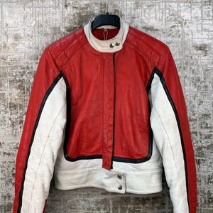 May include: A red and white leather jacket with black detailing. The jacket features a stand-up collar and quilted shoulder panels. The sleeves are white with black stripes, and it fastens with press studs.