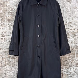 May include: A black, mid-length coat with a classic design. The coat has a collared neckline, button-down closure, and long sleeves with cuff details. It hangs from a black hanger against a textured background.