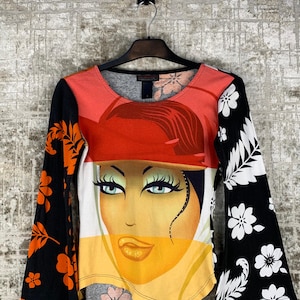Pode incluir: Camiseta de manga comprida com um gráfico do rosto de uma mulher. A camisa apresenta um design de bloco de cores com seções laranja, brancas e amarelas. As mangas são pretas com estampas florais laranja de um lado e brancas do outro.