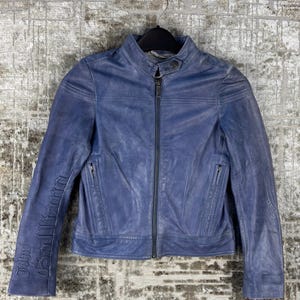Könnte beinhalten: Blaue Lederjacke mit Reißverschluss und Stehkragen. Die Jacke hat eine schmale Passform und trägt den Text "John Richmond" auf dem linken Ärmel. Die Jacke hängt an einem schwarzen Kleiderbügel.
