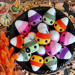 May include: A black basket filled with colorful, handmade crochet candy corn. The candy corn features a variety of colors, including orange, yellow, green, pink, and purple, with white tips and black embroidered details. The basket is surrounded by decorative fall elements.
