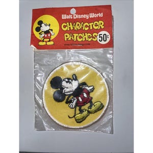 May include: A vintage Walt Disney World Character Patches patch featuring Mickey Mouse. The patch is round with a yellow background and a stitched border. The packaging includes the text "Walt Disney World Character Patches 50¢".