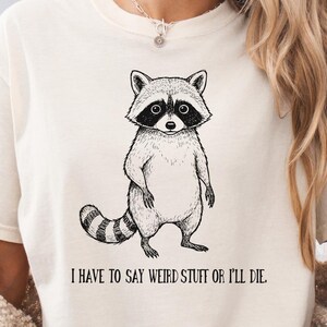 Puede incluir: Una camiseta de manga corta color crema con una ilustración en tinta negra de un mapache de pie. Debajo del mapache, el texto dice: "I HAVE TO SAY WEIRD STUFF OR I'LL DIE." La camiseta tiene un cuello redondo y un corte relajado.