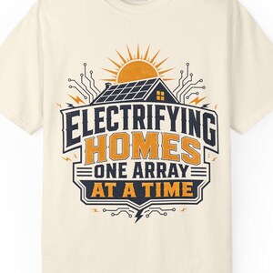 May include: A cream-colored short-sleeve t-shirt featuring a graphic of a house with solar panels, a sun, and circuit board patterns. The text reads: ELECTRIFYING HOMES ONE ARRAY AT A TIME in dark blue and orange lettering on the front of the shirt.