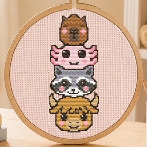 Könnte beinhalten: Ein runder Stickrahmen aus Holz mit einem Kreuzstichmuster, das einen vertikalen Stapel aus vier Tiergesichtern zeigt: ein braunes Wasserschwein, ein rosa Axolotl, ein grauer Waschbär und ein beiges Hochlandrind auf hellrosa Stoffgrund.