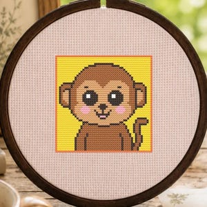 May include: A circular wooden embroidery hoop containing a cross-stitch pattern of a brown monkey with large black eyes and pink cheeks, centered on a bright yellow square background against a light pink fabric canvas with a visible grid texture.