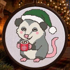 May include: A cross-stitch embroidery in a dark wooden hoop featuring a grey and white opossum wearing a green Santa hat with a white pom-pom. The opossum is holding a small red mug with a white snowflake design on its front.