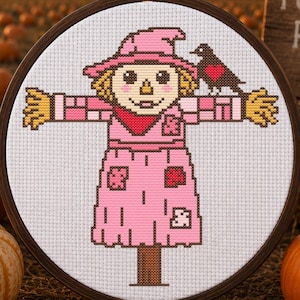 Può includere: Un ricamo a punto croce in un telaio circolare in legno raffigurante uno spaventapasseri con cappello rosa e vestiti rosa con toppe, in piedi con le braccia tese. Un piccolo uccello nero con un cuore rosso sul petto è posato sulla spalla.