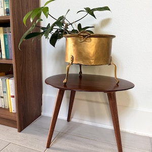 May include: A hammered brass pot with two side handles and three curved legs, containing a green leafy plant. The pot is positioned on a dark brown wooden side table with three tapered legs, standing near a wooden bookshelf filled with books.