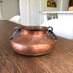 May include: A small, round, hammered copper bowl with a wide opening and a short, flared rim. The bowl features two dark, weathered metal ring handles attached to opposite sides of the rim, showing a natural patina and signs of surface wear.