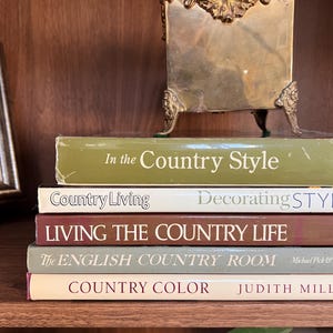 May include: A stack of books with titles like "In the Country Style" and "Country Living" sits on a wooden shelf. A decorative brass picture frame or holder with ornate detailing rests on top of the books. The books are various colors.