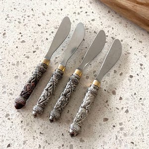 May include: Four butter knives with ornate, silver-colored handles and stainless steel blades. The handles feature a detailed, embossed design and are accented with gold-colored bands. A wooden cutting board is in the background.