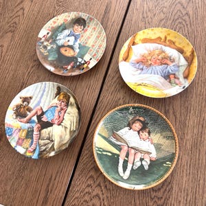 May include: Four decorative plates with children depicted in various scenes. One plate shows a boy playing a drum, another a girl in bed, a third a boy with a teddy bear, and the last two children reading in a hammock. The plates have a gold rim.