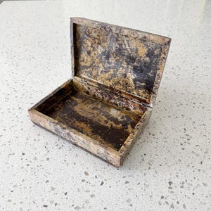 May include: A rectangular stone box with a hinged lid, featuring a polished finish and a mottled pattern of dark brown, black, and golden tan colors. The interior is open, revealing the same marbled stone texture throughout the base and lid.