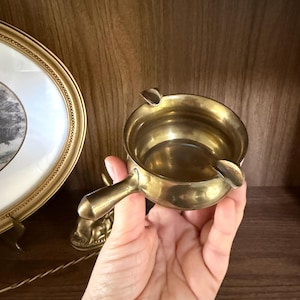 May include: A small, round, brass ashtray with a handle and two cigarette rests. The ashtray has a polished finish and is held in a hand. A gold-colored framed picture is in the background.