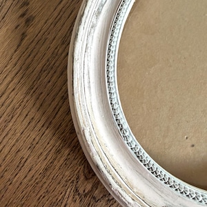 May include: A round picture frame with a distressed white painted finish over a light wood base. The frame features a raised, textured beaded inner border detail and a smooth, curved outer edge, surrounding a plain, light brown cardboard backing.