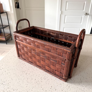 May include: A rectangular dark brown woven basket featuring a structured frame with a basket-weave pattern on the sides and base. The basket includes two arched handles attached to the shorter ends, secured with thin, wrapped material accents.