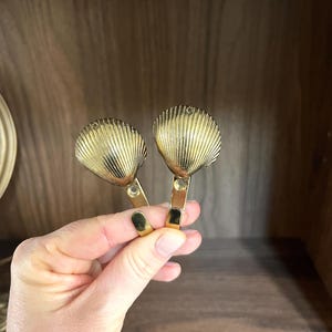 May include: Two gold-colored seashell-shaped hooks. The hooks are made of metal and have a shiny finish. The seashell design features detailed ridges. The hooks are held by a hand against a wooden background.