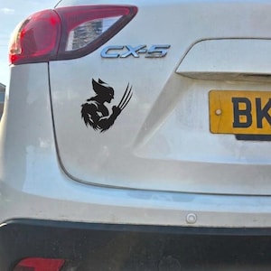 Könnte beinhalten: Ein schwarzer Vinyl-Aufkleber mit der Silhouette einer Figur, die drei scharfe Krallen an der Hand trägt, angebracht am Heck eines weißen Mazda CX-5. Das Motiv zeigt das Seitenprofil einer Gestalt mit spitzen Ohren und fellartiger Textur.
