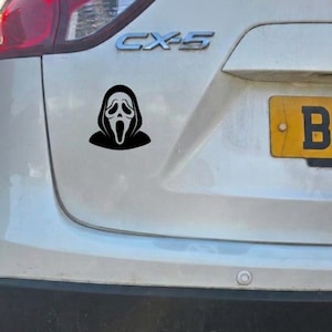 May include: A black vinyl decal featuring the iconic Ghostface mask from the Scream horror film franchise, applied to the white rear tailgate of a Mazda CX-5 vehicle, positioned between the taillight and the yellow license plate.