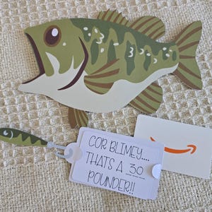 Puede incluir: Recorte de papel de un pez verde y marrón con la boca abierta, un pequeño señuelo de pesca, una etiqueta de regalo que dice "COR BLIMEY.... THAT'S A 30 POUNDER!!" y una tarjeta de regalo de Amazon.