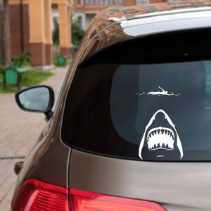 Könnte beinhalten: Weißer Vinyl-Autoaufkleber mit einem geöffneten Haifischmaul mit Zähnen und einem Schwimmer. Der Aufkleber ist auf der Heckscheibe eines Autos angebracht und erzeugt ein humorvolles Design.