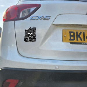 May include: A black vinyl decal applied to the rear of a white vehicle. The decal features a stylised graphic design of a stone castle structure integrated with a skull motif, positioned below the silver CX-5 emblem and above the rear bumper.