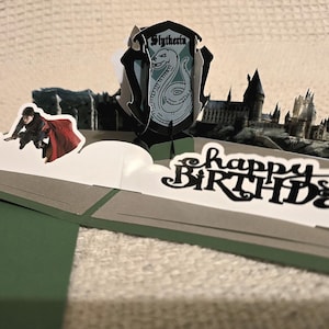 May include: A pop-up greeting card featuring a Slytherin house crest, a figure flying on a broomstick, a golden snitch, and a silhouette of a castle. The card includes the text "happy BIRTHDAY" in black lettering on a white background.