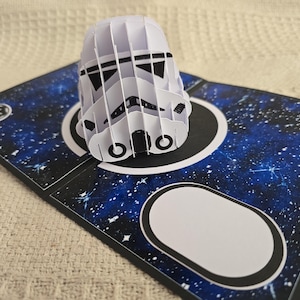 May include: A white, 3D paper Stormtrooper helmet sits atop a card with a blue galaxy design. The card features a black border, white circles, and star-like details. The card has a white oval shape.
