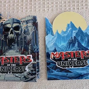Puede incluir: Dos tarjetas decorativas con el logo "Masters of the Universe". Una tarjeta representa una entrada de castillo en forma de calavera, mientras que la otra muestra una cordillera con un castillo y una luna amarilla. El logo es rojo y blanco.